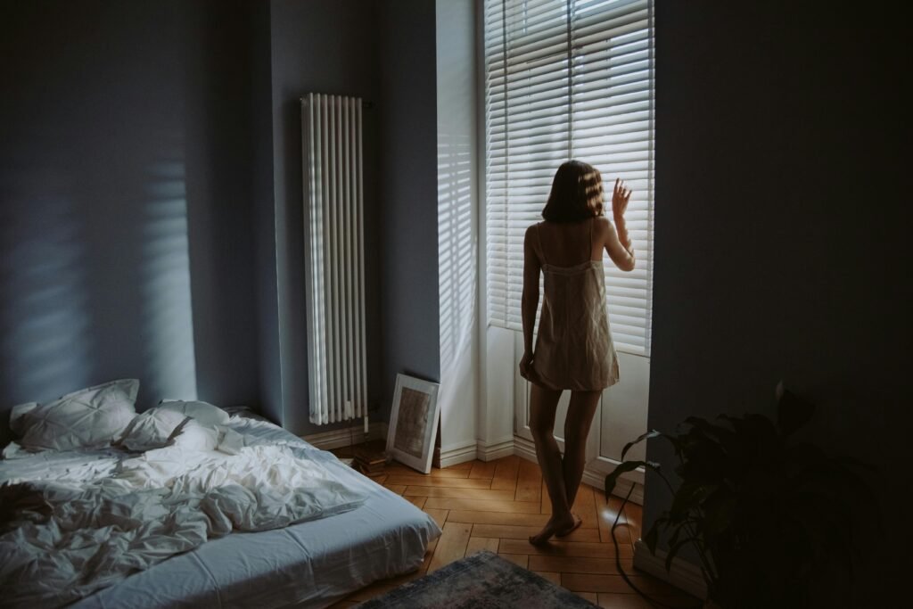 A woman in a bedroom stands by window blinds, casting shadows in soft morning light.